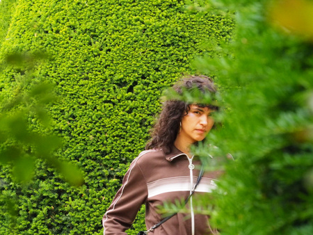 A woman in a brown track jacket framed by lush green garden foliage